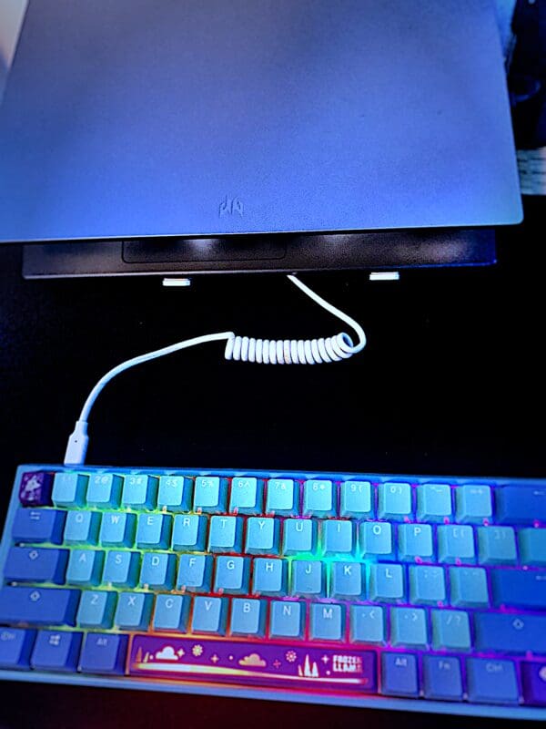 Colorful backlit mechanical keyboard with a USB 3.2 Male-A to Male-C Coil Cord - SS 10GBPS connected to a closed laptop in the background, on a dark desk.