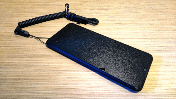 Black smartphone with a High Performance Coiled Lanyard Safety Tether attached, lying on a wooden surface.