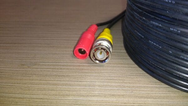Red and yellow RCA audio connectors and a coaxial Video Security Camera Wire with Connectors for CCTV Camera DVR Surveillance System end on a wooden surface, with a coil of cable in the background.